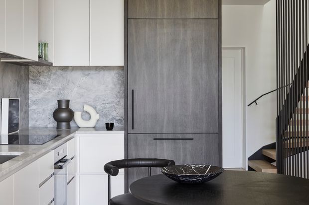 Contemporary open-plan kitchen viewed beside a staircase, featuring white cabinetry, a grey marble-look splashback and benchtop, a tall dark timber-look storage wall, and a round dark dining table with black chairs. Vertical black stair balustrades and timber stair treads add architectural definition.