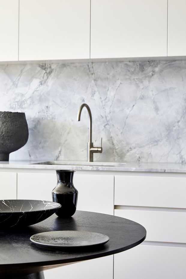 Close-up view of a refined contemporary kitchen with white flat-panel cabinetry, a dramatic pale veined stone splashback and matching benchtop, and a slim brushed-metal gooseneck tap. A dark round dining or breakfast table occupies the foreground, styled with black ceramic vessels and shallow plates.