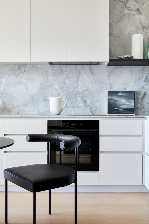 A refined contemporary kitchen featuring flat-panel white cabinetry, a black built-in oven, pale timber flooring, and a dramatic grey-white veined stone backsplash and benchtop. A black leather dining chair and partial round table occupy the foreground, while a small framed artwork, ceramic vessel, and open shelf add restrained decorative detail.