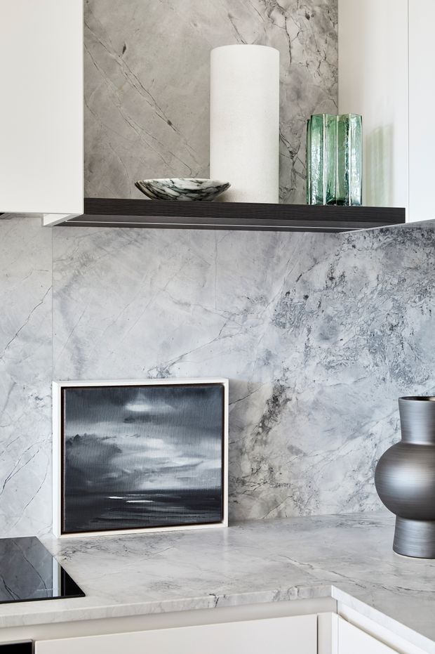 Close-up of a refined kitchen or built-in display area featuring dramatic grey-white veined natural stone across the wall and benchtop, white cabinetry, and a dark floating shelf. Minimal sculptural decor includes a white cylindrical vessel, a black-and-white bowl, translucent green glass objects, framed monochrome artwork, and a dark metallic vase.
