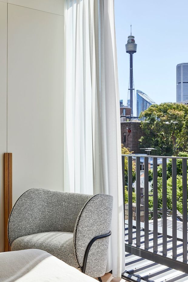 A bright contemporary interior corner with a textured upholstered lounge chair beside full-height sheer curtains and a large window or balcony opening. A dark vertical railing frames the balcony, with a clear urban skyline visible outside, including a prominent communications tower. The room features pale built-in wall panelling, warm timber trim, and soft neutral furnishings.
