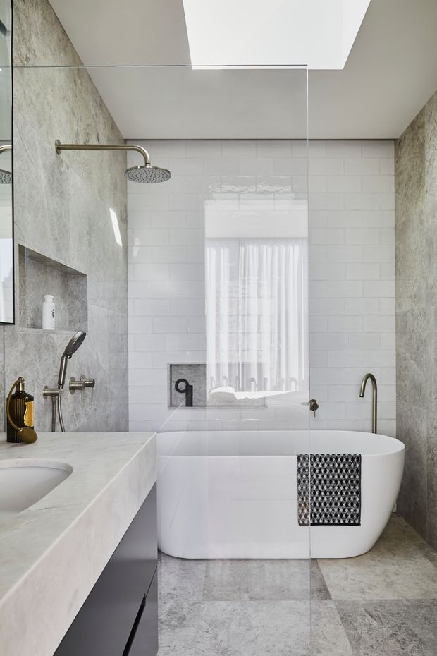 A bright contemporary bathroom with a freestanding white bathtub, wall-mounted shower, floating stone vanity, and large-format grey stone surfaces. White subway tiles line the rear wall, while a frameless glass shower screen and a skylight enhance the clean, airy feel. Brass-toned plumbing fixtures and a patterned towel provide subtle contrast.