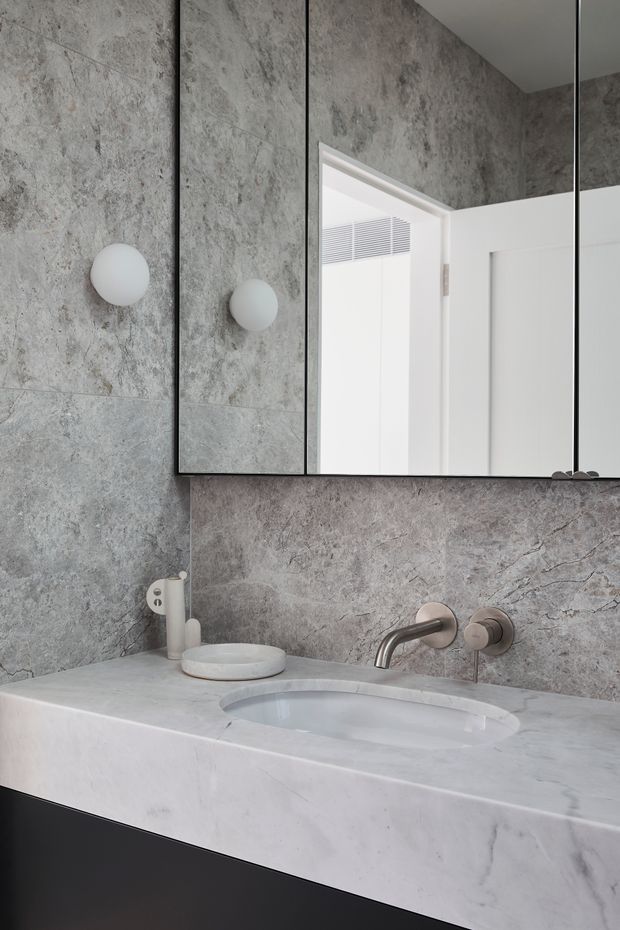 A refined contemporary bathroom vanity featuring grey stone-look wall tiles, a pale marble or engineered-stone benchtop with an integrated oval basin, wall-mounted brushed metal tapware, and dark under-vanity cabinetry. A large mirrored cabinet spans the wall above the vanity, reflecting a bright white doorway and ceiling vent. The space has a restrained, monochromatic palette and clean built-in detailing.
