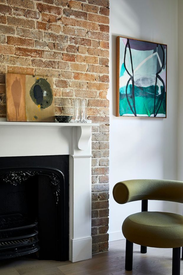 A close-up interior vignette featuring a traditional white-painted fireplace with ornate black cast-iron insert, exposed red brickwork, timber flooring, framed abstract artwork, and a curved olive-green upholstered armchair. The space blends period architectural details with contemporary furnishings and art.