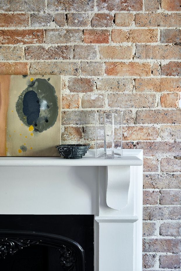 Close-up interior detail of a traditional white-painted fireplace mantel set against an exposed, weathered red and cream brick wall. The mantel supports a small abstract artwork, dark decorative bowl, and clear glass vessels, while the lower portion reveals ornate black fireplace detailing.