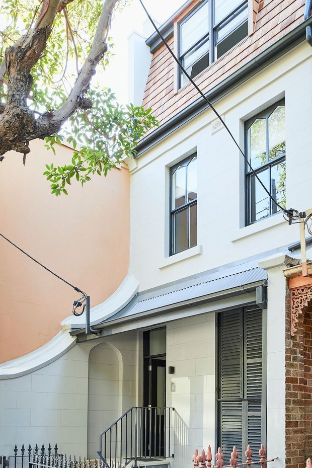 Exterior view of a narrow, multi-storey urban residence with a white rendered masonry facade, dark-framed windows, upper-level timber shingle cladding, corrugated metal awning, arched entry recess, black louvred shutters, and decorative metal fencing. Mature tree branches and adjoining heritage-style brickwork frame the compact courtyard-like frontage.