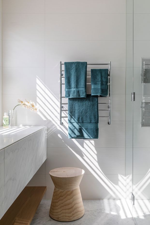 Bright contemporary bathroom featuring large pale wall tiles, a marble-look floating vanity, chrome heated towel rail with deep teal towels, glass shower enclosure, and a sculptural light timber stool. Strong sunlight creates striped shadows across the walls and floor.