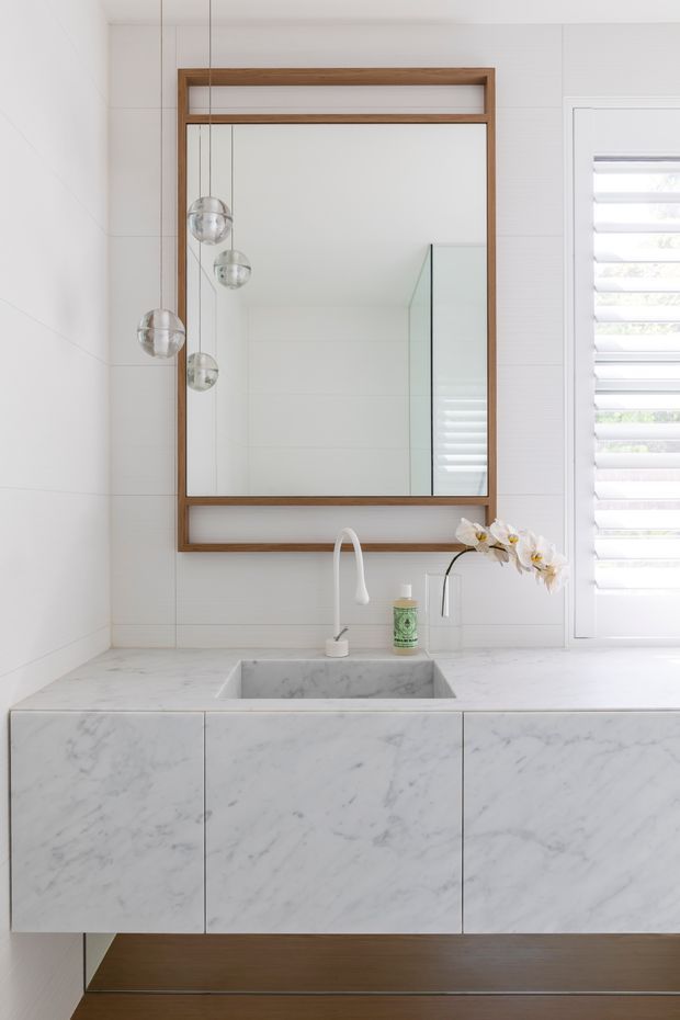 Bright, minimalist bathroom vanity featuring a floating marble-clad cabinet with integrated rectangular basin, white curved faucet, large timber-framed mirror, white tiled walls, and a louvred window. Globe pendant lights and a small orchid add subtle decorative detail.