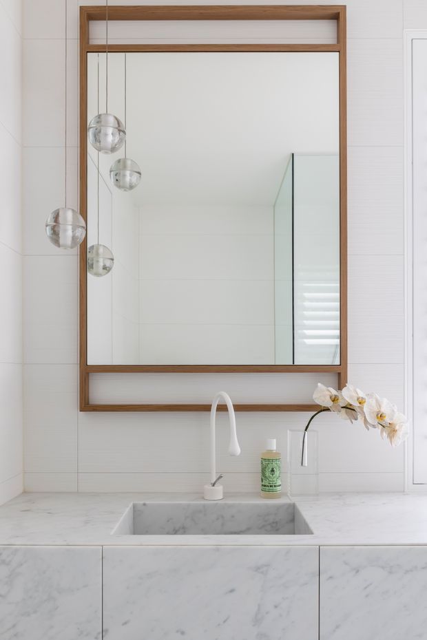 Bright, refined bathroom vanity featuring a large rectangular mirror in a warm timber frame, white tiled walls, a pale marble benchtop and integrated rectangular basin. A white curved faucet, wall-mounted tap, pendant globe lights reflected in the mirror, and a small orchid arrangement complete the space.