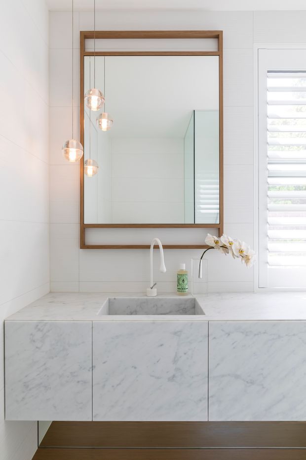 Bright contemporary bathroom vanity with a floating white marble cabinet and integrated rectangular stone basin. The white tiled walls are paired with a large framed timber mirror, suspended globe pendant lights, a curved white tap, and white plantation shutters. A warm timber floor and minimalist detailing create a clean, refined interior.