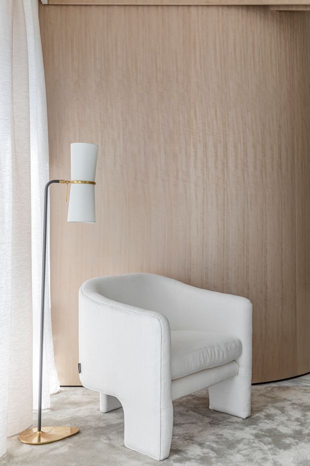 A refined interior vignette featuring a sculptural white upholstered armchair, a slim floor lamp with a brass-gold base and accents, and a pale timber wall panel behind them. Soft sheer curtains and a neutral patterned carpet create a calm, understated setting.