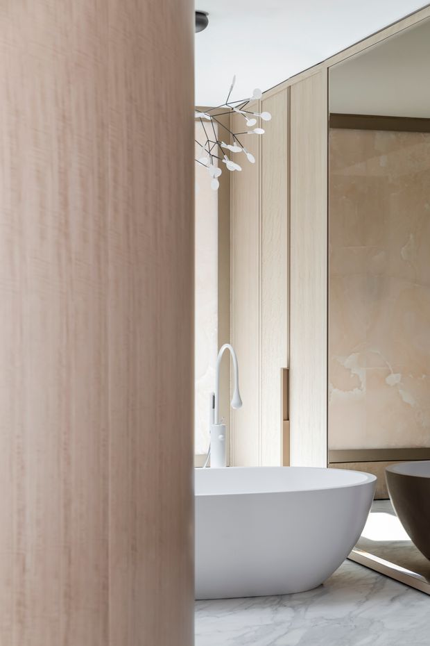 A refined contemporary bathroom featuring a freestanding white bathtub, pale timber wall cabinetry or panelling, a light marble-look floor, and a sculptural white pendant light. A large rounded timber-clad partition partially screens the view, while a warm beige textured feature wall and vanity area appear in the background. The palette is soft, neutral, and luxurious with abundant natural light.
