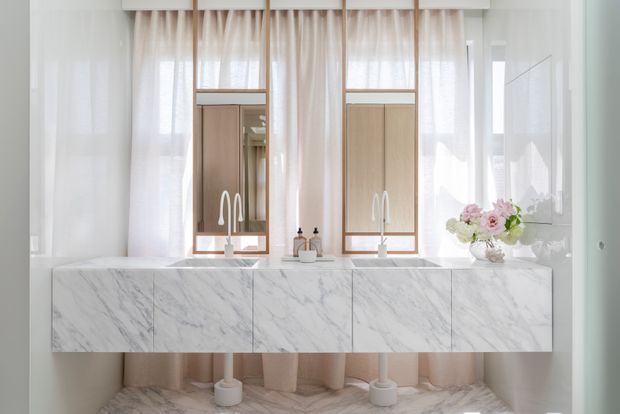 Bright, refined bathroom vanity featuring a long floating white marble vanity with integrated basins and sculptural chrome faucets. Two framed mirrored medicine cabinets are set within pale timber-toned joinery, with sheer curtains, soft neutral walls, and floral accents creating an airy, elegant interior.