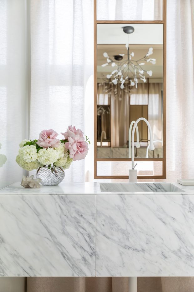 Bright, refined bathroom vanity vignette featuring a large white-and-grey marble vanity with integrated rectangular basin, a warm timber-framed mirror, sheer curtains, and soft floral styling. The mirror reflects a sculptural branching pendant light and additional cabinetry, while a curved white tap rises from the countertop.