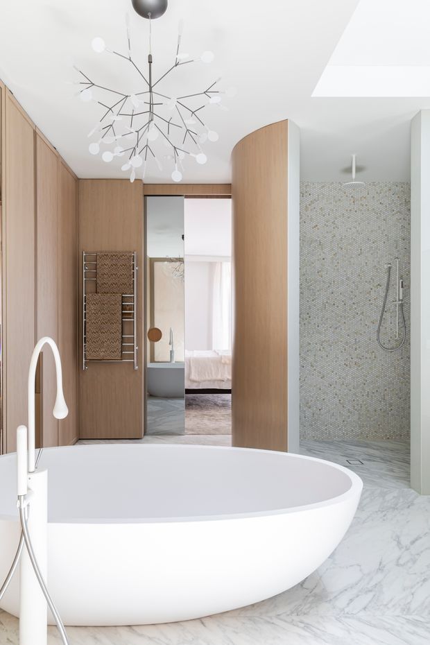 Bright contemporary bathroom featuring a sculptural freestanding white bathtub, pale marble-look flooring, a walk-in shower with small mosaic tile walls, warm timber wall panelling and cabinetry, and a large branching pendant light. A bedroom is visible through the open doorway beyond the bathroom.