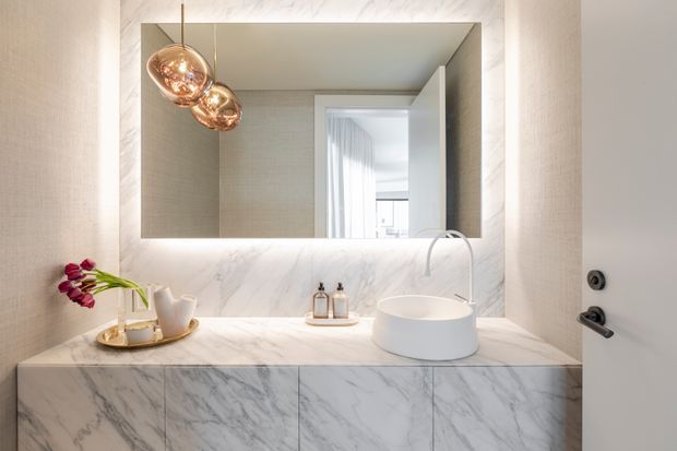 Contemporary bathroom vanity with a white vessel basin, brushed metal tapware, marble-look wall and cabinetry surfaces, and a large illuminated mirror. Warm copper pendant lights and minimal styling add a refined, luxurious feel. An open door and reflected corridor are visible in the mirror.