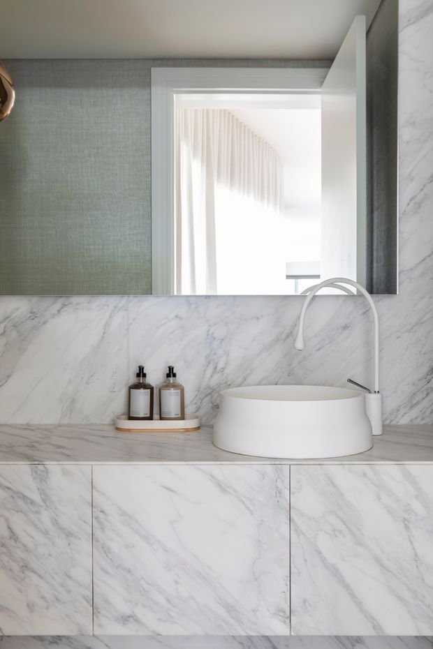 Close-up view of a refined bathroom vanity featuring a white vessel basin, curved white tapware, and extensive white-grey veined marble across the benchtop, backsplash, and vanity cabinetry. A large mirror reflects a bright adjoining space with sheer curtains, while a muted green textured wall panel adds subtle contrast.
