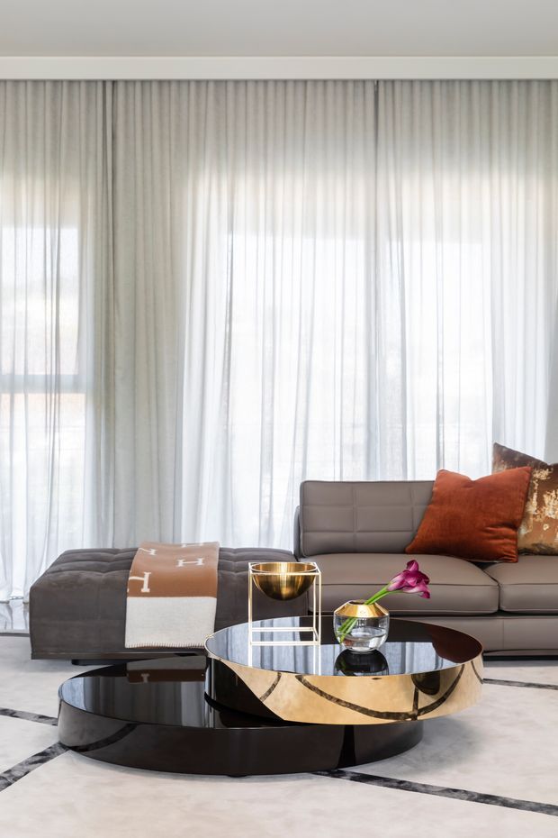 Bright, sophisticated living room featuring a large grey sectional sofa in front of full-height sheer curtains. Layered round coffee tables combine a glossy black base with a metallic gold-toned upper table, while rust-orange and patterned cushions add warmth. A pale stone or marble-look floor with dark geometric inlay lines completes the refined contemporary setting.