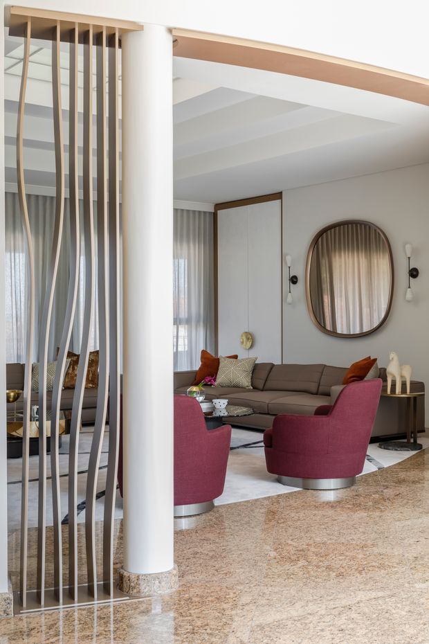 A refined living room with a curved brown sofa, burgundy lounge chairs, a large oval timber-framed mirror, sculptural wall sconces, and sheer drapery. A decorative vertical screen of slim timber-toned slats separates the seating area from an adjacent space. The room features a polished mottled stone floor, white walls and ceiling, curved architectural details, and warm wood accents.