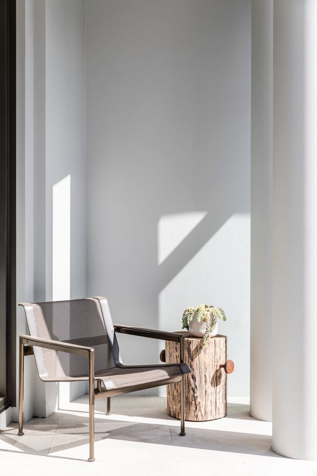 A minimalist sunlit architectural interior with a modern mesh lounge chair beside a rustic natural wood stump side table. Tall white walls and cylindrical columns create a sculptural, gallery-like space, with strong geometric sunlight and shadows across the walls and pale floor. A small potted plant adds an organic accent.