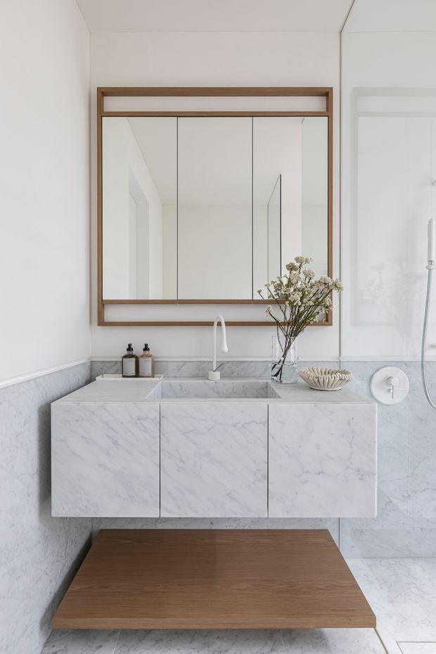 A bright, refined bathroom vanity area featuring a floating white marble vanity with integrated basin and white tapware, pale marble wall and floor finishes, and a large three-panel mirrored cabinet framed in warm timber. A timber floating shelf or platform sits below the vanity, with restrained decorative accessories and a glass shower visible at right.