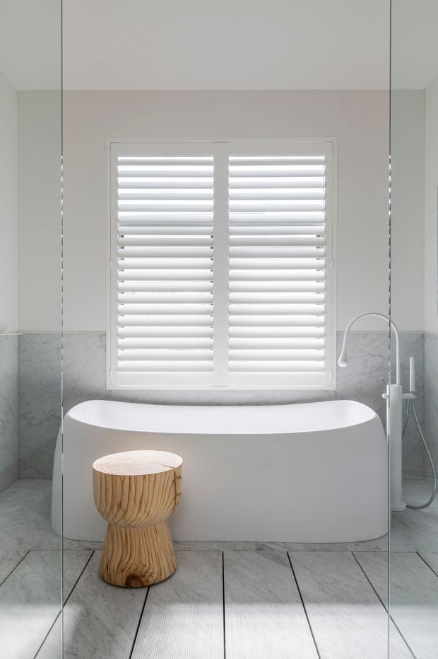 Bright contemporary bathroom featuring a freestanding white bathtub beneath a white shuttered window, pale marble-look wall tile, light stone-look floor tiles, and a sculptural natural timber stool. Frameless glass shower panels are visible at both sides.