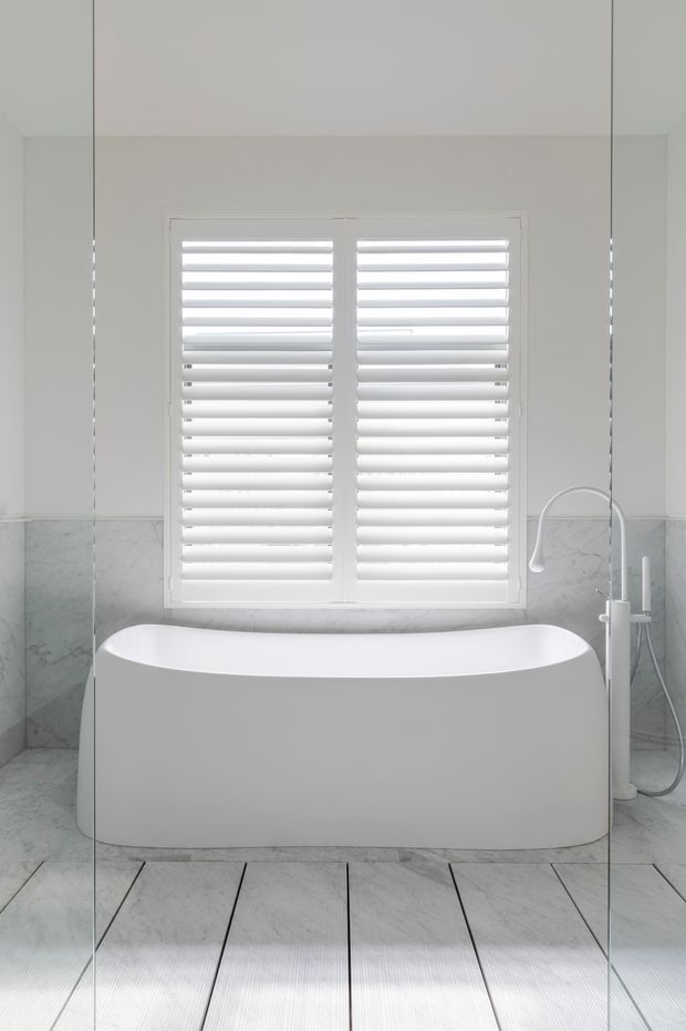 Bright, minimalist bathroom featuring a freestanding white bathtub positioned beneath a large window with white plantation shutters. Pale marble-look wall and floor tiles create a clean, light-filled setting, with a white freestanding bath and shower fixture visible at right.