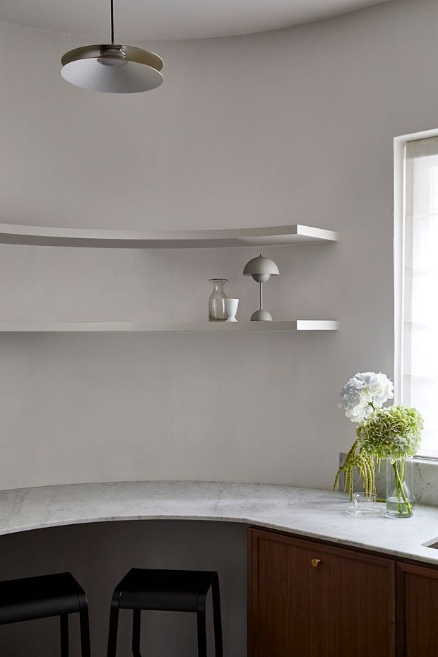 A refined kitchen corner with a curved pale stone benchtop, dark timber cabinetry, black stools, integrated open shelving, and soft grey walls. A sculptural pendant light and small decorative objects sit above the counter, while daylight enters through the window beside a vase of flowers. The overall composition is minimal and understated with a warm material contrast between the stone, timber, and black furniture.
