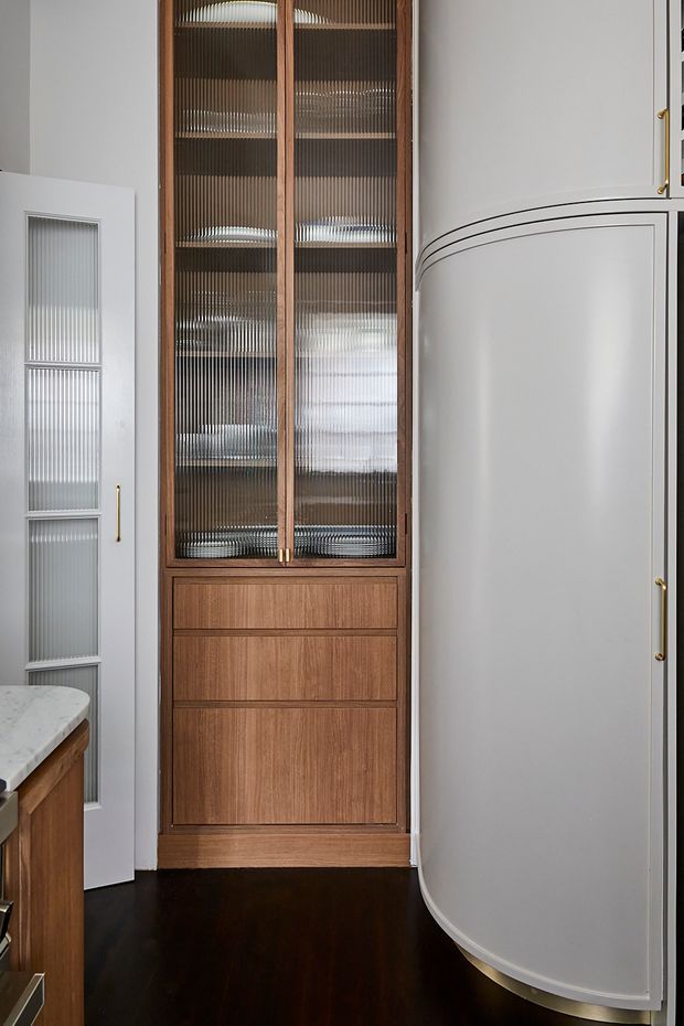 A refined interior joinery detail showing a tall built-in timber cabinet with ribbed translucent glass doors and lower drawers, framed by white painted cabinetry. A curved white cabinet door occupies the right side, while a small portion of a stone benchtop and timber cabinetry is visible at lower left. The dark timber floor and warm wood tones create a sophisticated, tailored fit-out.