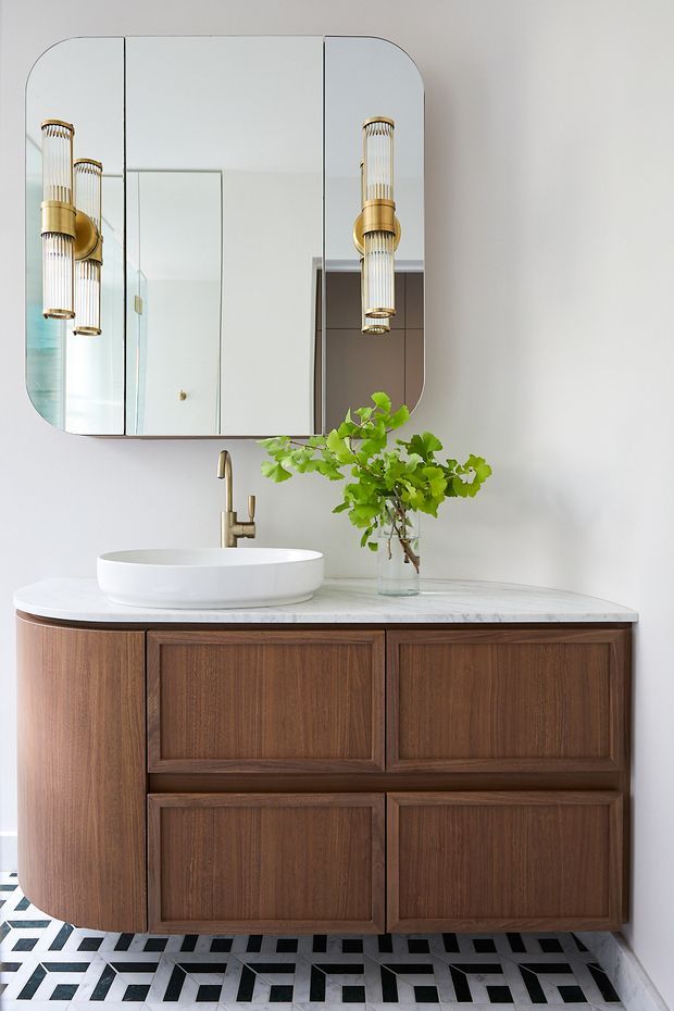A refined bathroom vanity with a warm dark timber floating cabinet, white stone benchtop, white vessel basin, and brushed brass tapware. A large rounded-corner mirrored cabinet is mounted above, accompanied by two decorative vertical brass-and-glass wall lights. The space features a bold geometric black-and-white tiled floor and minimalist white walls, with a small leafy plant adding an organic accent.