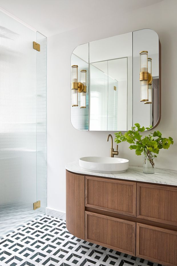 Bright, refined bathroom featuring a floating dark timber vanity with framed panel cabinetry, a white vessel basin, brushed brass tapware, a large rounded-corner mirrored cabinet, and decorative brass-and-glass wall sconces. A fluted glass shower enclosure and bold geometric black-and-white floor tiles add texture and pattern.