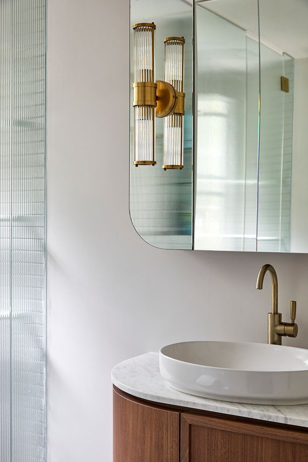 A refined bathroom vanity featuring a white vessel basin on a white marble countertop, warm timber cabinetry, and a brushed brass wall-mounted tap. A rounded-corner mirrored cabinet and matching brass-and-glass wall sconce create a luxurious focal point, while vertically fluted translucent wall glazing adds texture and diffused light.