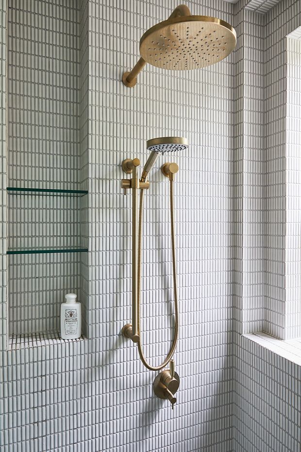 A compact shower enclosure lined with small white vertically ridged tiles. The shower features brushed brass fixtures, including a large round overhead rain shower, handheld shower, controls, and wall-mounted outlets. A recessed tiled niche with dark glass shelves and a small bottle is visible on the left, while a tiled ledge and bright window opening appear on the right.