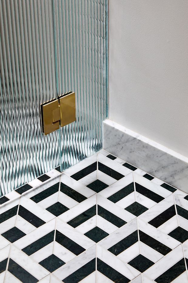 Close-up architectural detail of a refined bathroom or powder room, featuring a geometric black-and-white marble tiled floor, matching marble skirting, a textured vertically patterned glass wall, and a gold-toned wall-mounted flush plate. The composition emphasizes bold geometric surfaces and luxurious material contrast.