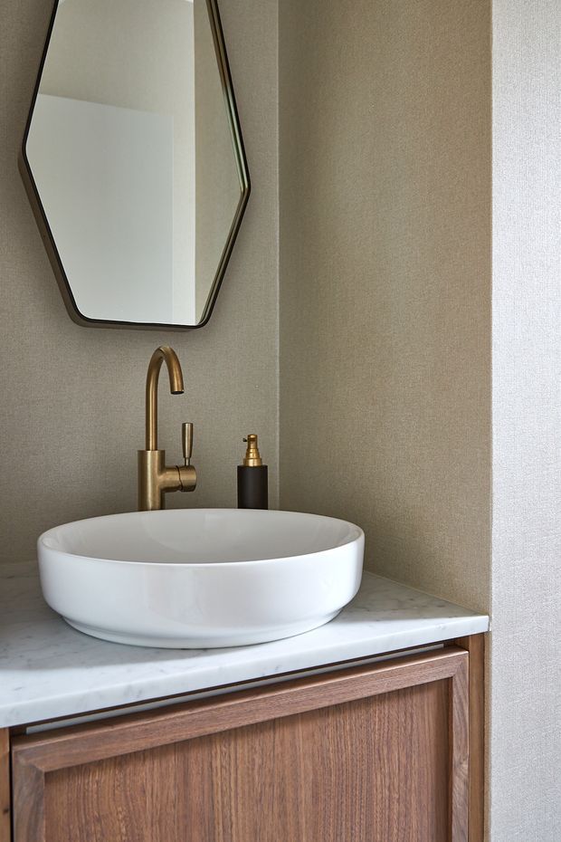 Close-up view of a refined bathroom vanity featuring a white vessel basin on a white marble countertop, warm brass tapware, timber cabinetry, textured neutral walls, and an organically faceted wall mirror.