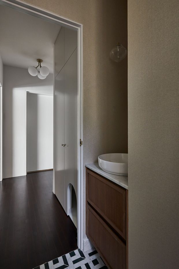 A narrow, refined bathroom or powder room viewed from the hallway, featuring a floating dark timber vanity with integrated drawers, a white vessel basin, pale stone benchtop, textured taupe walls, and a small wall-mounted glass light. Bold black-and-white geometric floor tiles define the threshold, while dark timber flooring and a white-paneled doorway lead into the adjacent hall. A spherical multi-light pendant is visible in the adjoining space.