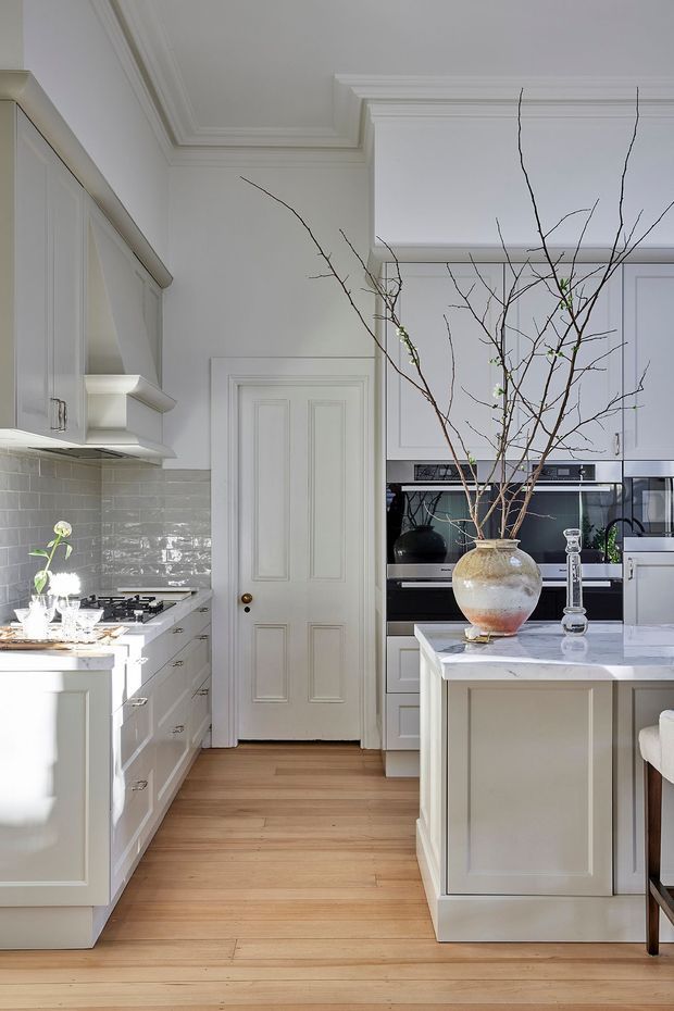 Bright, refined kitchen with white Shaker-style cabinetry, grey glazed subway-tile splashback, pale timber flooring, and a marble-topped central island. Integrated black appliances are set into the cabinetry at the rear, while a decorative vase with branches forms a focal point on the island. The room features ornate ceiling cornices and detailed white trim around the internal door.