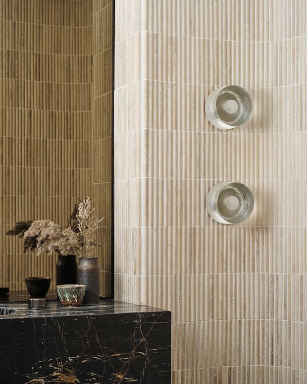 Close-up of a refined bathroom or vanity area featuring vertically fluted beige and cream wall tiles, a dramatic black marble ledge with gold veining, and two round metallic wall sconces. Decorative ceramic vessels and dried botanicals sit on the ledge, creating a warm, textural and luxurious composition.