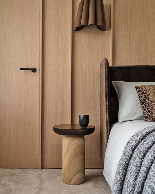 A warm, refined bedroom detail featuring full-height light timber wall panelling and a concealed or flush timber door with a dark handle. A sculptural dark wood side table with a small black vessel sits beside an upholstered dark headboard, white bedding, and a textured grey throw. The palette is earthy and tonal, with subtle sculptural wall decor above the bedside table.
