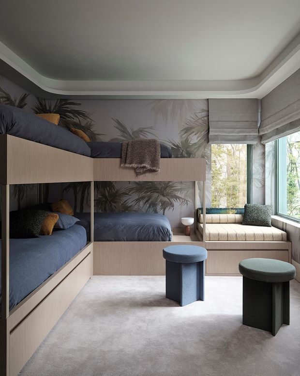 A compact, thoughtfully designed bedroom with built-in bunk beds and integrated storage in pale timber cabinetry. The room features muted blue bedding, a soft grey carpet, palm-patterned wallpaper, a built-in window seat, upholstered stools, and large windows with Roman blinds. Rounded ceiling detailing and curved furniture edges create a refined, contemporary feel.