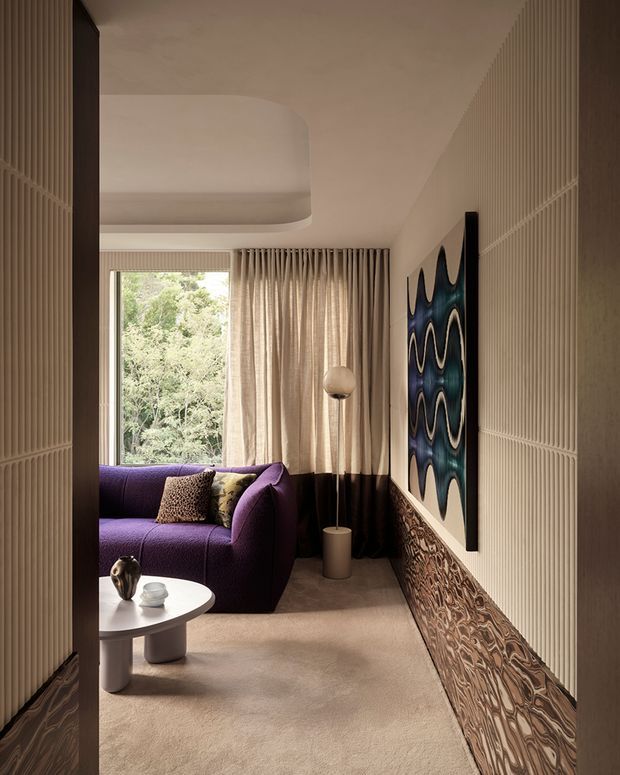 A compact, refined living room viewed from the entry, featuring a deep purple curved sofa, small round white coffee table, sculptural floor lamp, full-height beige curtains, and a large abstract artwork. The room has softly rounded ceiling detailing, textured vertical wall panelling, an ornate patterned lower wall treatment, and a warm neutral carpet. Natural greenery is visible through the window.