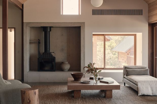 Warm, refined living room with a built-in concrete fireplace surround and black wood-burning stove, timber-lined ceiling, large timber-framed windows, and a low solid timber coffee table. A neutral sofa and chaise lounge sit on a textured rug, with natural light and a view toward a neighbouring timber structure outside.