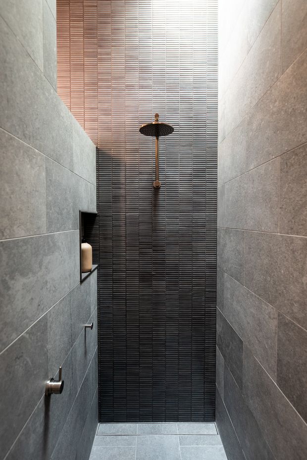 A narrow, contemporary walk-in shower enclosed by large-format grey wall tiles. The rear wall features dark, finely ribbed tiles creating a strong vertical focal surface, with a ceiling-mounted round rainfall shower and matching dark hardware. A recessed wall niche holds a bottle, and the floor is finished in coordinating grey tile. Natural light filters through horizontal blinds or slatted glazing above.
