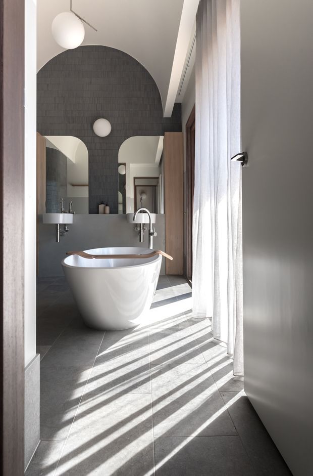 A refined contemporary bathroom viewed through a doorway, featuring a freestanding white bathtub, wall-mounted basin, large arched mirror, dark textured tiled feature wall, grey floor tiles, timber cabinetry, and sheer full-height curtains casting strong diagonal shadows across the floor. Sculptural spherical pendant and wall lights add a minimalist decorative element.
