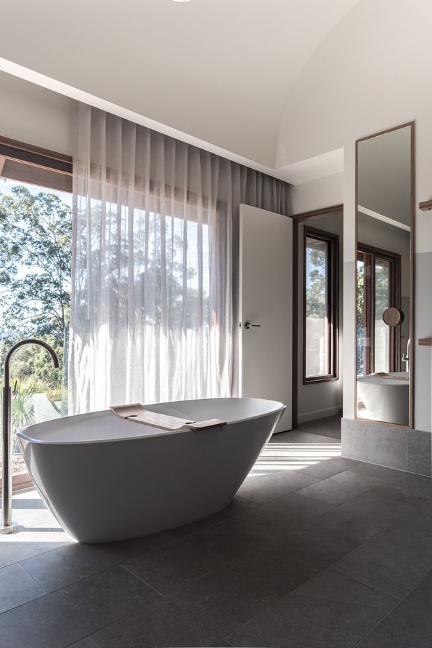 A bright, contemporary bathroom featuring a freestanding white sculptural bathtub, dark grey tiled flooring, sheer full-height curtains, dark timber-trimmed windows and door, and a tall framed mirror. Natural light and views of trees enter through the glazing, creating a calm indoor-outdoor atmosphere.