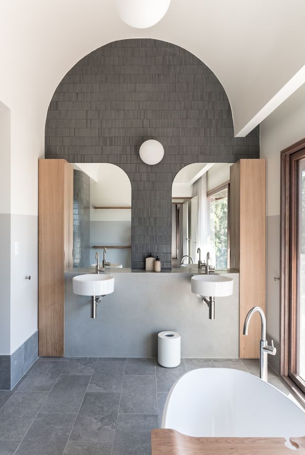 A contemporary bathroom featuring a freestanding white bathtub in the foreground, twin wall-mounted white basins, exposed chrome plumbing, and a large mirrored vanity wall. The central wall is clad in dark vertically ribbed tiles with a dramatic arched and curved profile, flanked by warm timber cabinetry panels. Large timber-framed windows bring in natural light, while grey floor tiles and two-tone painted walls complete the refined interior.