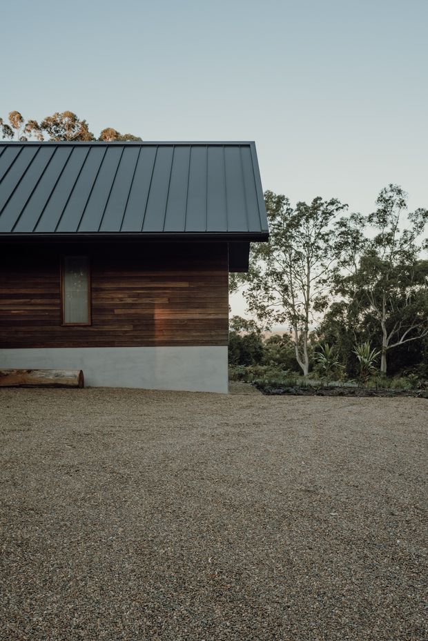 Exterior view of a compact contemporary building in a landscaped bush setting. The facade combines dark horizontal timber cladding with a pale concrete base, a narrow vertical window, deep dark eaves, and a steep standing-seam metal roof. A broad gravel forecourt occupies the foreground, with native-looking trees and planting behind the building.