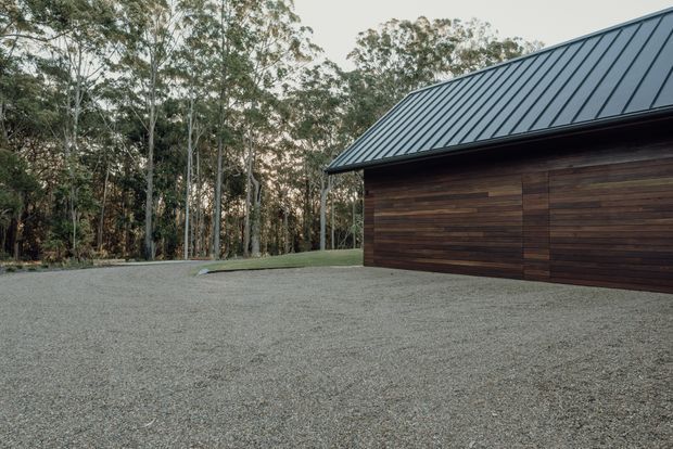 Exterior view of a simple contemporary building set within a wooded landscape. The structure has dark horizontal timber cladding, a standing-seam metal pitched roof, and a broad gravel forecourt. Tall trees and lawn surround the building.