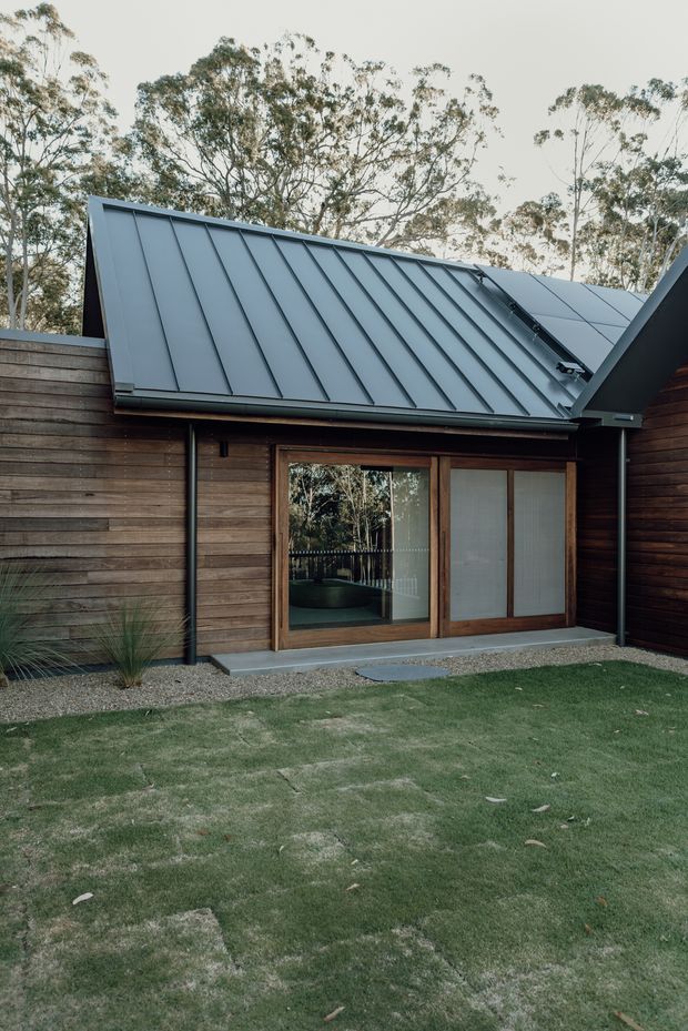 Exterior view of a compact contemporary building set within a landscaped garden. The facade combines horizontal dark timber cladding with large timber-framed glazed sliding doors, while a steep asymmetrical roof is finished in dark grey standing-seam metal. Gravel borders, lawn, and mature trees surround the structure.
