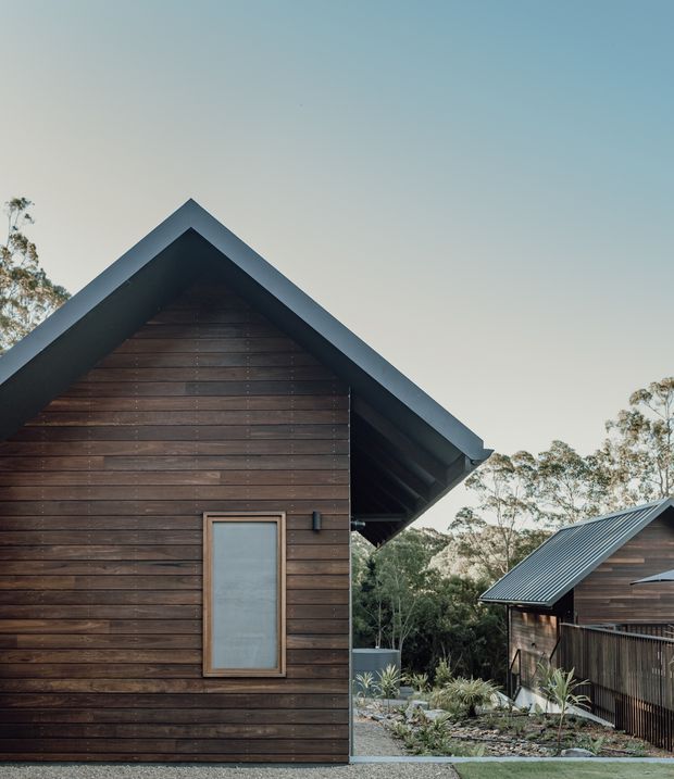 Exterior architectural photograph of a compact dark timber-clad building with a steep gabled roof and deep roof overhang. A second matching timber structure is visible beyond a landscaped gravel and planting area, set among trees. The composition emphasizes the weathered horizontal cladding, dark metal roof edges, and simple cabin-like forms.